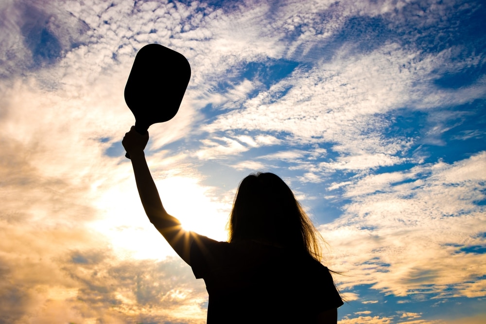 A back shadow image of a woman holding a pickleball paddle under a blue sky